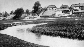 Fotografia czarno-biała przedstawia krajobraz wiejski. Na pierwszym planie wijąca się rzeka, w oddali drewniane zabudowania.