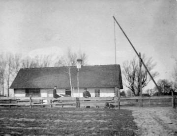 Fotografia czarno-biała przedstawia dwóch mężczyzn przy płocie, na tle wiejskiej chaty. Po prawej stronie żuraw studzienny. 