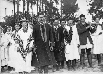 Fotografia czarno-biała przedstawia grupę ludzi ubranych w biłgorajski strój ludowy. Drugi mężczyzna od prawej strony trzyma skrzypce.