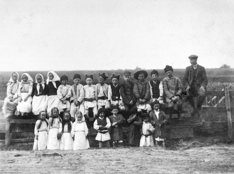 Fotografia czarno-biała przedstawia grupę dzieci w strojach ludowych. Dzieci ustawione w dwóch rzędach, przy drewnianym ogrodzeniu, na tle łąki.