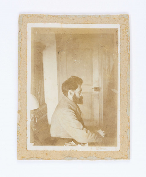 Stefan Żeromski siedzący na tle drzwi z silnym zarostem, z wąsami i brodą w marynarce sfotografowany z profilu.