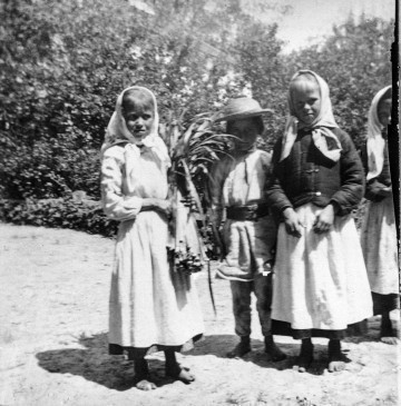 Fotografia czarno-biała przedstawia czworo dzieci ubranych w stroje ludowe. Dziewczynka na pierwszym planie trzyma roślinę. W tle gęste zarośla. 