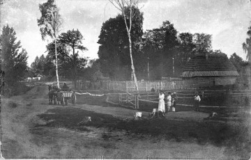 Fotografia czarno-biała przedstawia krajobraz wiejski. Na pierwszym planie droga, po której jedzie wóz zaprzęgnięty w konie oraz grupa dzieci bawiących się przy zagrodzie. W tle widoczne drewniane zabudowania wiejskie. 
