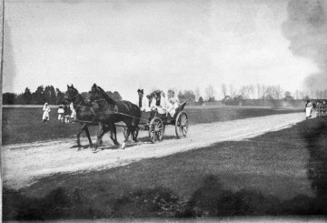 Fotografia plenerowa czarno-biała przedstawia grupę w strojach ludowych jadącą na wozie zaprzęgniętym w dwa konie. W oddali kilka osób idzie pieszo, a po lewej stronie kilka osób stoi na polu i obserwuje przejeżdżający wóz. W tle krajobraz wiejski z zabudowaniami.  