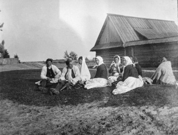 Fotografia czarno-biała przedstawia grupę osób, która siedzi na tle budynku drewnianego.