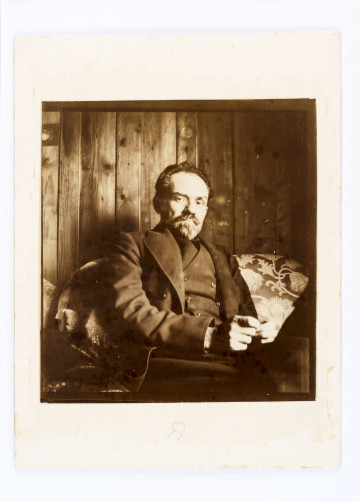 Stefan Żeromski w marynarce, z brodą i wasami, siedzi na kanapie na tle ściany z desek.  Ręce trzyma na nogach blisko siebie.