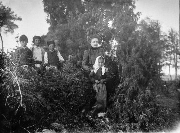 Fotografia czarno-biała przedstawia grupę dzieci z opiekunką na tle drzew i krzaków.