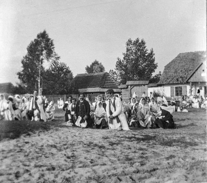 Fotografia czarno-biała przedstawia grupę mieszkańców wsi ubranych w stroje ludowe, którzy na klęczkach oczekują na obrzęd poświęcenia pokarmów. W oddali widoczne drewniane zabudowania wiejskie.