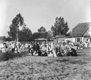 Fotografia czarno-biała przedstawia grupę mieszkańców wsi ubranych w stroje ludowe, którzy na klęczkach oczekują na obrzęd poświęcenia pokarmów. W oddali widoczne drewniane zabudowania wiejskie.