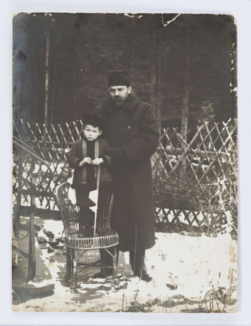 Mały Adaś w kożuszku oparty na lasce stoi na krześle, obok stoi Stefan Żeromski w długim palcie i baraniej czapce.