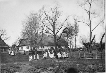 Fotografia czarno-biała przedstawia krajobraz wiejski. Grupa w strojach ludowych zgromadzona wokół drewnianej zabudowy.