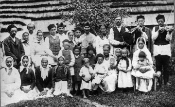 Fotografia czarno-biała przedstawia grupę w stojach ludowych. W pierwszym rzędzie kobiety i dzieci, w drugim rzędzie stoją kobiety i mężczyźni. Po prawej stronie trzej mężczyźni z instrumentami muzycznymi. W tle widoczna strzecha ze słomy. 