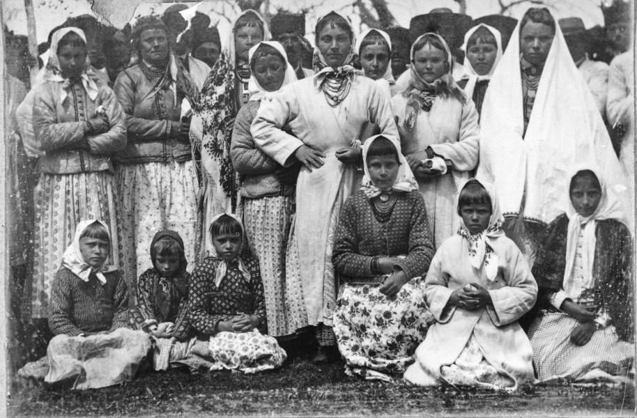 Fotografia czarno-biała przedstawia grupę w strojach ludowych. W pierwszym rzędzie dzieci siedzące na trawie, w drugim rzedzie stoją kobiety, za nimi mężczyźni. 