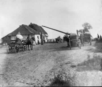 Fotografia czarno-biała przedstawia wiejską drogę, po której jadą wozy zaprzężone w konie. Po lewej stronie drewniany budynek ze strzechą. 