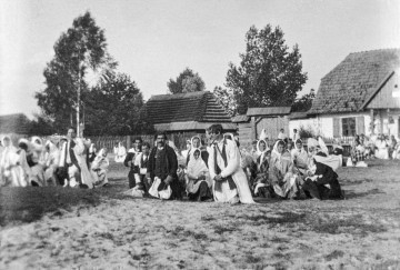 Fotografia czarno-biała przedstawia grupę mieszkańców wsi ubranych w stroje ludowe, którzy na klęczkach oczekują na obrzęd poświęcenia pokarmów. W oddali widoczne drewniane zabudowania wiejskie. 