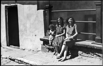 Fotografia czarnobiała dawnej dzielnicy żydowskiej. Widać dwie młode kobiety i dziecko siedzące na murku pod balustradą na mostku przy ulicy Zamkowej nad Bramą Zasraną (ob. nieistniejącą) przy chodniku wyłożonym w jodełkę płytkami. Kobieta po prawej stronie uśmiechnięta, zwrócona do widza, z lekko kręconymi ciemnymi włosami, z lewą nogą założoną na prawej, w jednolitej kolorystycznie sukience i szmacianych podniszczonych butach, trzyma w rękach zwinięte w rulon papiery. Kobieta w środku, z bosymi stopami, ubrana w biało-czarną sukienkę we wzory geometryczne, w rozpuszczonych włosach, uśmiechnięta, mrużąca lewe oko, lewą rękę z pierścionkiem na palcu serdecznym ma ułożoną na prawym udzie, prawą zaś trzyma siedzącego obok małego chłopca. Dziecko z kręconymi włosami, ze wzrokiem zwróconym w lewo, w jasnej koszulce, trzyma w rękach kawałki jedzenia (chleba?); na nogach ma sandały. Po lewej stronie fotografii wejście do otynkowanego i pobielonego budynku. W lewym dolnym rogu fragment brukowanej ulicy i rynsztok.