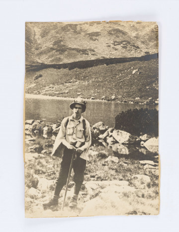 Fotografia przedstawia Adama Żeromskiego stojącego na tle stawu, na drugim planie zbocze góry. Ubrany w  kapeluszu, bluzce z kieszeniami, spodnie narciarskiem buty oraz skarpety wywijane na wierzch. Oparty na laseczce, z plecakiem. Rękawy bluzy rozpiete, luźno zwisające.