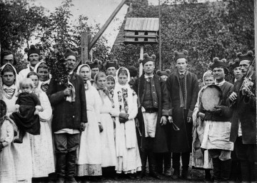 Fotografia czarno-biała przedstawia grupę mężczyzn, kobiet i dzieci w strojach ludowych podczas uroczystości weselnych. Mężczyzna stojący po lewej stronie trzyma rózgę weselną, a mężczyźni stojący po prawej stronie - instrumenty muzyczne. 