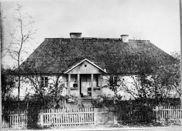 Fotografia czarno-biała przedstawia drewniany dom ogrodzony płotkiem. W ganku stoi kobieta ubrana na czarno, towarzyszy jej czarny pies. 