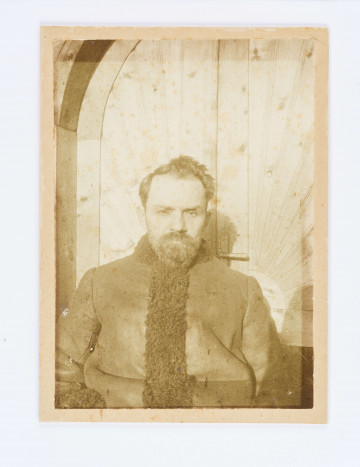 Pisarz z odkrytą głową, z brodą i wąsami siedzi na tle drzwi, w baranim kożuchu lamowanym futrem.