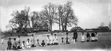 Fotografia czarno-biała. Grupa osób na tle drzew oraz zabudowań drewnianych, m.in. cerkwi.