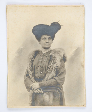 Maria Radziwiłowicz (1/2 postaci) w kapeluszu, oparta o plecione krzesło.