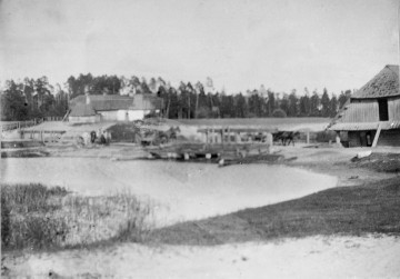 Fotografia czarno-biała przedstawia dwa drewniane zabudowania wiejskie usytuowane przy brzegu zbiornika wodnego. W tle widoczna grupa ludzi i konie. W oddali rząd wysokich drzew. 