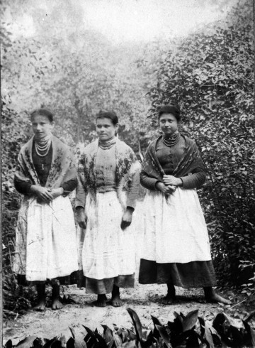 Fotografia czarno-biała przedstawia trzy kobiety na tle drzew i krzaków. Są ubrane w biłogorajskie stroje ludowe.