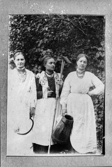 Fotografia czarno-biała przedstawia trzy kobiety na tle drzewa. W rękach trzymają narzędzia, m.in kosę i grabie.