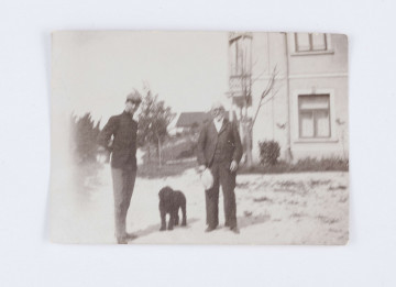 Fotografia przedstawia Emila Trembińskiego z nieznanym mężczyzną. Emil stoi po lewej stronie, ubrany jest w czarny surdut oraz szare spodnie, a na głowie znajduje się szary kaszkiet. Ręce trzyma za plecami. Po prawej stronie stoi starszy mężczyzna ubrany w ciemną marynarkę, spodnie, kamizelkę oraz białą koszulę. W prawej ręce trzyma biały kapelusz. Między mężczyznami widać czarnego psa. W tle, po prawej stronie, widać fragment  murowanego, jasnego, piętrowego budynku. Po lewej stronie znajduje się kilka drzew i  krzaków. W oddali widać zarysy pojedynczej chaty. Na odwrocie znajduje się odręczny napis: „Nałęczów/ Emil Trembiński/ R. 1900”.