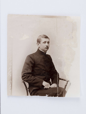 Fotografia przedstawia młodego Emila Trembińskiego siedzącego na krześle. Młodzieniec ubrany jest w czarny surdut zapięty aż pod szyję, białą koszulę oraz ciemne spodnie. Wzdłuż lewej krawędzi widać nieczytelny, biały napis. Prawa cześć fotografii mocno pogięta. Na odwrocie znajduje się napis wykonany ołówkiem: 
