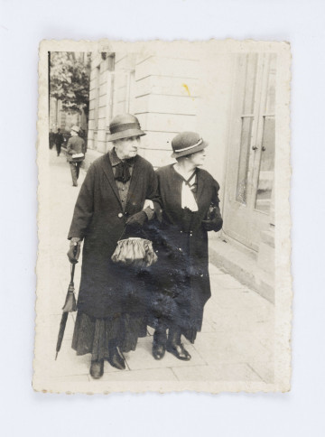 Fotografia przedstawia Oktawie Głowacką oraz Janinę Juszkiewiczową. Pani Oktawia znajduje się po lewej stronie, ubrana jest w czarny płaszcz, szarą koszulę, czarną apaszkę, ciemną spódnicę oraz kapelusz. W prawej ręce trzyma parasolkę, a w lewej trzyma torebkę. Po prawej stronie widać panią Janinę. Ubrana jest w czarny płaszcz oraz białą apaszkę. W lewej dłoni, pod pachą, trzyma torebkę. Panie idą brukowanym chodnikiem, po prawej stronie widać biały, budynek z oszklonymi drzwiami. W głębi zdjęcia widać spacerującego mężczyznę. Na odwrocie znajduje się odręczny napis wykonany ołówkiem: „ p. Oktawia Głowacka/ i Janina Juszkiewiczowa „Emilka”.