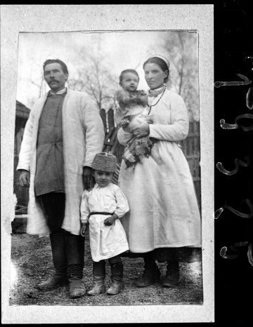 Fotografia czarno-biała przedstawia rodzinę w strojach biłgorajskich. Postacie stoją na tle drewnianego płotu. Od lewej strony: ojciec, kilkuletni syn i matka z niemowlęciem na ręku. 