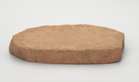 Płyta szlifierska - ujęcie z boku; Płyta szlifierska wykonana z płaskiego fragmentu czerwono-pomarańczowego piaskowca. Na jednym z końców widoczne są ślady przygotowania prostej powierzchni, co sugeruje, że płyta powstała poprzez podział większej bryły przy wykorzystaniu naturalnego podziału skały osadowej na warstwy. Na stronie wierzchniej płyta ma wyraźne ślady użytkowe w postaci zagładzeń, od spodu nie posiada żadnych śladów.