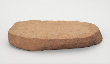 Płyta szlifierska - ujęcie z boku; Płyta szlifierska wykonana z płaskiego fragmentu czerwono-pomarańczowego piaskowca. Na jednym z końców widoczne są ślady przygotowania prostej powierzchni, co sugeruje, że płyta powstała poprzez podział większej bryły przy wykorzystaniu naturalnego podziału skały osadowej na warstwy. Na stronie wierzchniej płyta ma wyraźne ślady użytkowe w postaci zagładzeń, od spodu nie posiada żadnych śladów.
