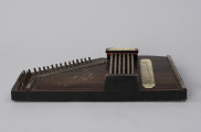 Cytra klawiszowa (autoharp) - ujęcie z trzeciego boku; Cytra klawiszowa (autoharp) z płaskim pudłem rezonansowym wykonanym z drewna polichromowanego o kształcie przypominającym trapez z jednym ściętym rogiem. Wzdłuż najkrótszego boku oraz boku odchodzącego od niego pod kontem rozwartym, szereg metalowych kołków, do których przymocowanych jest 25, diatonicznie strojonych strun, ułożonych od najgrubszej – z dołu – do najcieńszej w górnej części instrumentu; ostatnie dwie są zerwane. Na prawo od kołków znajdują się dwie, zestawione pod kątem rozwartym, wąskie, metalowe blaszki z niewielkimi bolcami, stanowiące wsparcie i gwarantujące właściwy rozstaw strun. Podobna blaszka zamontowana jest wzdłuż dolnej krawędzi instrumentu. Struny przechodzą przez znajdujące się w niej rowki, zaginają pod kątem 90 stopni. Bezpośrednio nad strunami, prostopadle względem nich, znajduje się sześć metalowych prętów zamontowanych pomiędzy dwoma drewnianymi mostkami. Osadzone na sprężynach pręty wprawiane są w ruch przez grającego za pomocą znajdujących się w ich górnej części przycisków. Powoduje to dociśnięcie do struny mocowanych na prętach klawiszy z filcowymi podkładkami. Na jednym z mostków, na przykręconej za pomocą dwóch śrub plakietce z papieru welinowego, znajdują się oznaczenia akordów. Druga plakietka, z zapisem nut w formie literowej i na pięciolinii oraz ciągiem cyfr rzymskich i arabskich, umieszczona jest bezpośrednio na pudle rezonansowym, na prawo od mechanizmu klawiszowego. Cytra ozdobiona jest w prawej części kalkomanią.