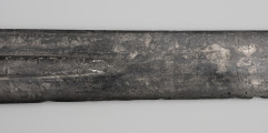 Miecz jednoręczny typu XII (Oakeshott) - detal; Na końcu fragmentu inskrypcji grecka omega oraz symbol w formie liścia o zakreskowanym polu połączonym kreską z rąbem, przed nią rozeta w formie kółka z promieniami zakończonymi „perełkami” odczytywana jako znak solarny.