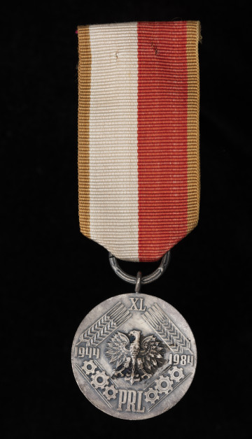 Medal 40-lecia Polski Ludowej - ujęcie z przodu; Odznaka Medalu 40-lecia Polski Ludowej w postaci srebrzonego, oksydowanego krążka ze wstążką i przylutowanym uszkiem. Na awersie medalu w środku umieszczono wizerunek orła, a po obu jego stronach daty: 1944 i 1984. Nad orłem znajduje się rzymska cyfra: XL, a pod orłem napis: PRL. Daty połączone są z rzymską liczbą stylizowanymi kłosami zboża, a z napisem kołami zębatymi. Wszystkie elementy są wypukłe. Wstążka medalu jest szerokości 32 mm, składająca się z dwóch pionowych pasków w środku w kolorze białym i czerwonym o szerokości 12 mm każdy i dwóch pasków po bokach o szerokości 4 mm koloru złotego. Wstążka zawieszona na kółku w formie rózg liktorskich o średn. 16 mm, przechodzącym przez uszko medalu.