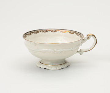 Filiżanka do herbaty - ujęcie z przodu; Duża filiżanka z białej porcelany dekorowana złoceniami. Czarka filiżanki okrągła w przekroju poziomym, szeroka na wysokości wylewu zwęża się w kierunku dna. Poniżej wylewu złocony ornament w formie ukośnego szrafowania ograniczonego od góry poziomą dookolną linią i od dołu ułożonymi na przemian C-kształtnymi elementami nawiązującymi do uproszczonego ornamentu małżowinowo-chrząstkowego. Na zewnętrznej ściance czarki oraz na stopce delikatne dookolne pasmo ornamentu plastycznego w formie spłaszczonych esownic i rozciągniętego motywu małżowinowo-chrząstkowego. Nawiązuje do niego również forma uszka ozdobionego pasmem złocenia na stronie grzbietowej, które przechodzi w cienką strużkę podkreślającą plastyczność ślimacznicy na profilu uszka, tuż przy łączeniu górnej jego części z czarką.