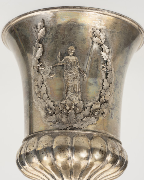 Puchar Hartwiga - detal; Ujęcie górnej części srebrnego pucharu. Czara w kształcie odwróconego dzwonu ozdobiona jest w dolnej części niskim puklowanym koszyczkiem z wybrzuszeniem, oddzielonym od płaszcza czaszy wąskim pierścieniem z ornamentem perełkowym. Gładką, rozszerzającą się ku górze powierzchnię czary zdobi dekoracja nakładkowa personifikacja sprawiedliwości - Temida z opaską na oczach, z wzniesionym mieczem w lewej i dwuszalkową wagą w prawej ręce. Postać ujęta w otwarty górą wieniec z liści dębowych z żołędziami (niektóre niemal pełnoplastyczne).