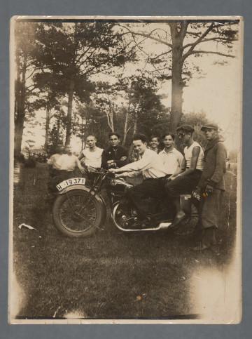 Chłopiec stojący najbardziej po lewej: figura poruszona. Dwie spośród sfotografowanych osób – na motorze, druga ma gogle. Nr rej. motoru: W 19371. Obraz z fotografii z przejaśnieniami w rogach. Krawędzie proste. Zdjęcie wywołane z niewielkimi marginesami, u góry i u dołu widoczne przed marginesem czarne paski, wynikające z wady wywołania. Jedna z pamiątek, jak możemy sądzić, Mani Żelaznej.