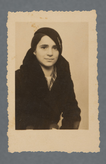 Monochromatyczna fotografia studyjna młodej kobiety - Mani Żelaznej - w berecie i płaszczu; popiersie; ujęta w półprofilu.Krawędzie ząbkowane. Zdjęcie wywołane z marginesem, nie jest on zupełnie równy.