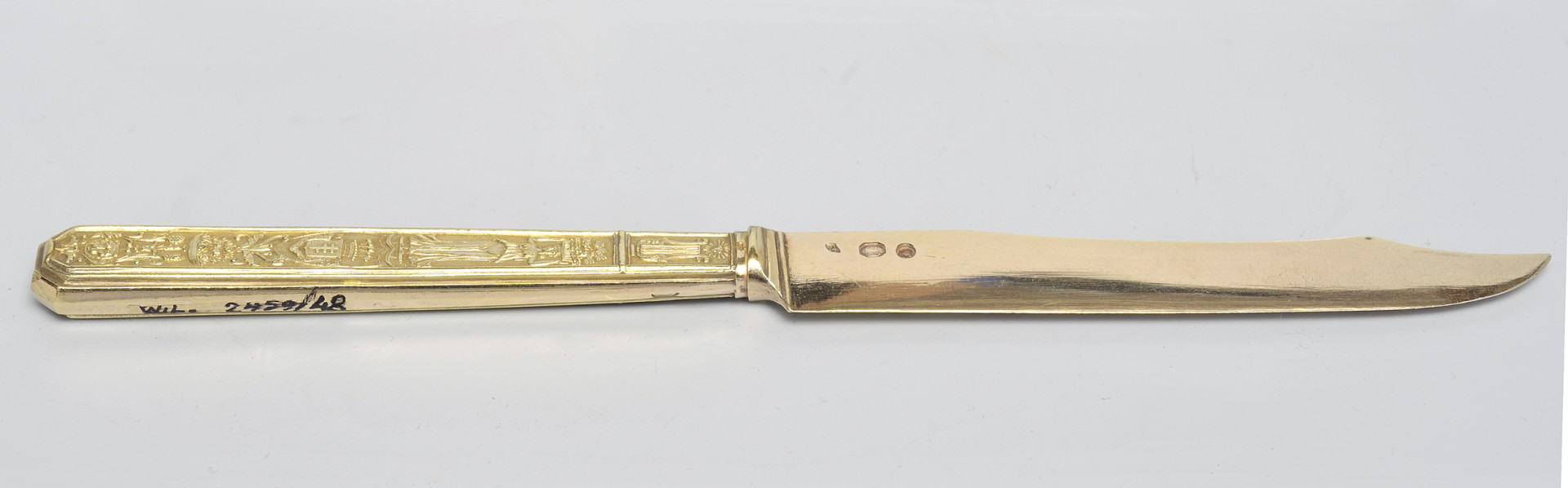 ujęcie 3/4 - strona z puncami na głowni. Trzy punce o różnych kształtach przy łączeniu głowni z rękojeścią. Ujęcie z widoczną krawędzią tnącą i szczytem ostrza skierowanym w prawo. Na dolnej krawędzi trzonka numer inwentarzowy.