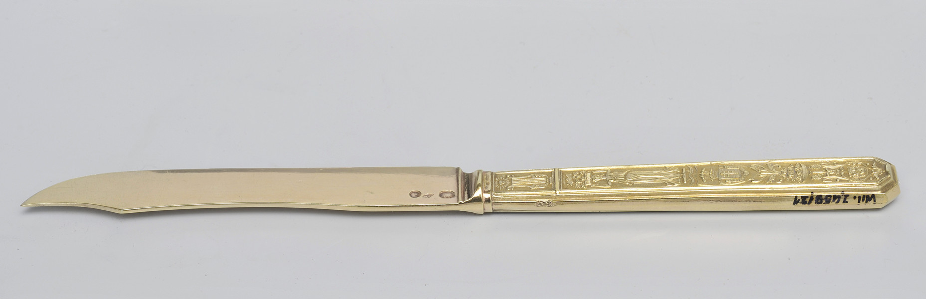 ujęcie 3/4 - strona z puncami na głowni. Trzy punce o różnych kształtach przy łączeniu głowni z rękojeścią. Ujęcie z widocznym grzbietem i szczytem ostrza skierowanym w lewo. Na końcu górnej krawędzi trzonka numer inwentarzowy.