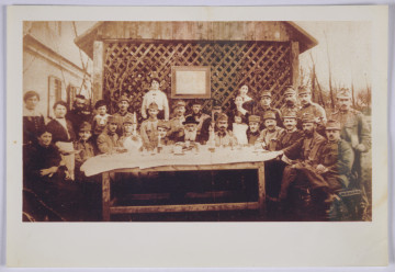 Fotografia rodzinna z austriackimi żołnierzami - Zdjęcie grupowe wykonane w czasie święta Pesach. Wtórna odbitka. Ozjasz Grossbart (siedzi pośrodku stołu), dziadek Emilii Leibel, zaprosił żołnierzy austriackich pochodzenia żydowskiego na wspólne obchody. Jeden z żołnierzy ma na rękawie opaskę z czerwonym krzyżem. Na zdjęciu widoczne także kobiety: jedna z nich, w fartuchu, trzyma talerz, jakby zatrzymała się na moment zrobienia zdjęcia w trakcie nakrywania do stołu, inna kobieta, w ciemnej sukni, siedząca, trzyma chochlę. W tle widoczny obraz, o którym wiemy z relacji, że był podobizną cesarza Franciszka Józefa. Uroczystość zorganizowano na zewnątrz, stół ustawiono na tle drewnianej ściany konstrukcji w formie pergoli.