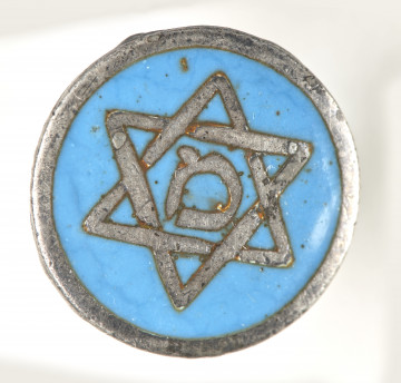 Metalowa okrągła odznaka, emaliowana w kolorze niebieskim. W środkowej części odznaki widoczna jest gwiazda sześcioramienna (heksagram) o barwie szarej, w jej centralnym punkcie umieszczona została hebrajska litera מ (pol. m), również w odcieniu szarym. Na rewersie przylutowano zapięcie w postaci śruby. 