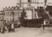 zbliżenie na dolny kadr fotografii. Na pierwszym planie po lewej kiosk z gazetami oraz kilku przechodniów zmierzających ku ulicy Andrijejewski Zjazd opadającej stromo wzdłuż muru cerkwi