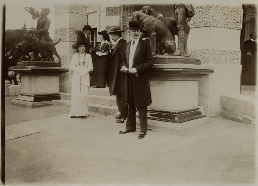 Na fotografii widoczni są ludzie stojący przed pałacem w Antoninach. Na pierwszym planie stoi mężczyzna w meloniku, obok niego mężczyzna w kapeluszu, a obok nich kobieta w białej sukni w kapeluszu. 