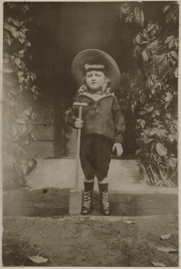 Na zdjęciu znajduje się chłopiec stojący przez drzwiami pałacu, trzyma w ręku szpadel.
