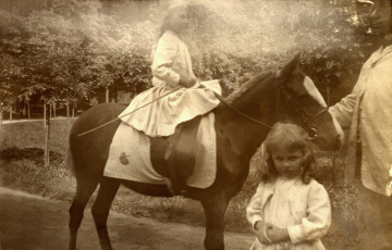 Na pierwszym planie fotografii znajduje się dziecko, tuż za nim na kucu trzymając lejce i szpicrutę dziewczynka, obok pracownik ordynacji trzymający częściowo kuca za uzdę.